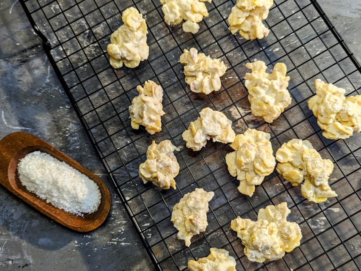 Ein schwarzes Gitter auf einem grauen Steintisch mit dem Raffaello-Knusper. Links daneben ein paar echte Raffaellos und eine Holzschippe mit Kokosflocken.