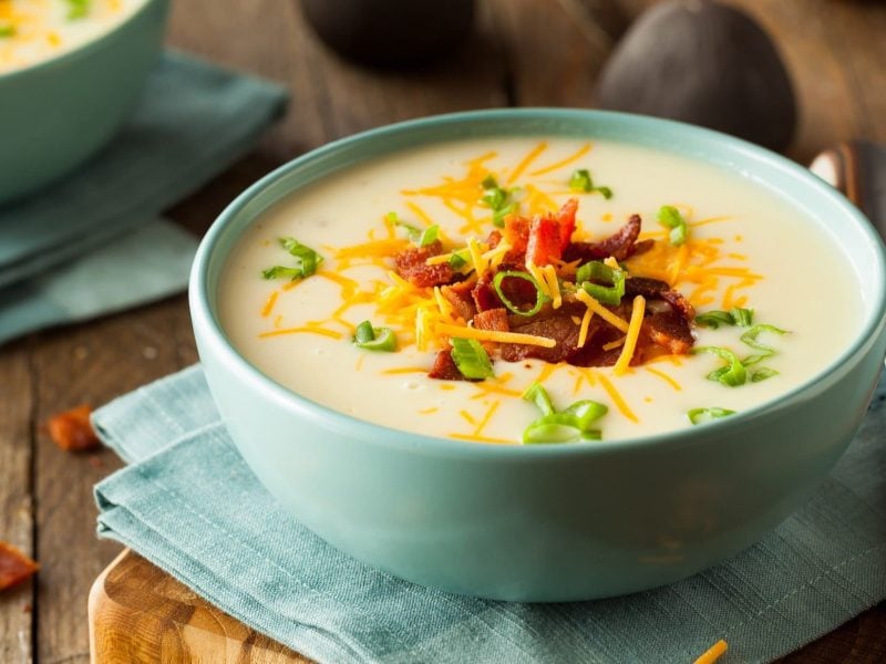 Draufsicht, schräg: Eine Raclette-Suppe mit Speck in einer hellblauen Schüssel. Sie steht auf einem Tuch aus Leinen. Daneben liegt ein Suppenlöffel.