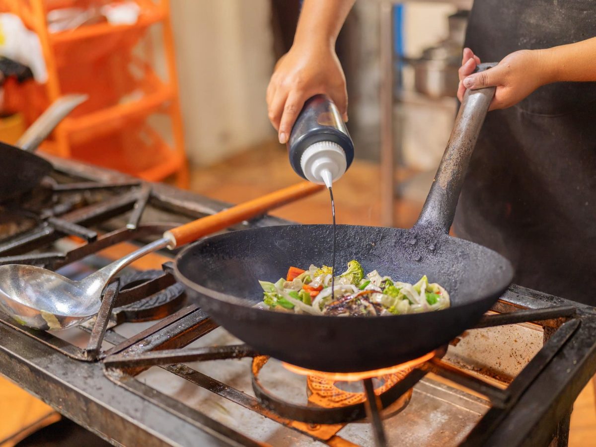 Eine Hand hält eine Wok-Pfanne, in der er über offenem Feuer Gemüse gart. In der anderen Hand hält er einen Plastikfleisch mit einer der drei besten Sojasaucen.