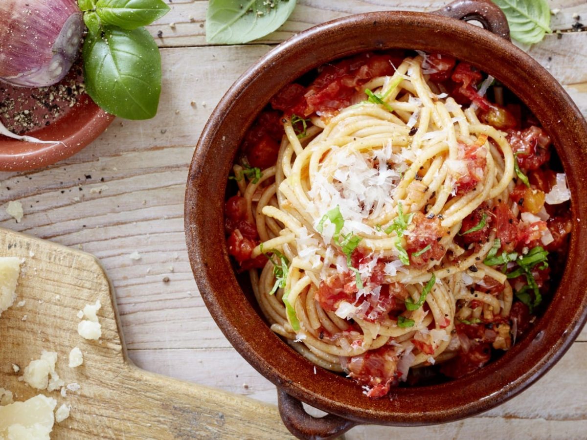 Spaghetti mit Chili in einem braunen Topf auf hölzernem Hintergrund. Links davon ein Brett mit Parmesan und links oben Schalotten, Knoblauch, Basilikum und Pfeffer.