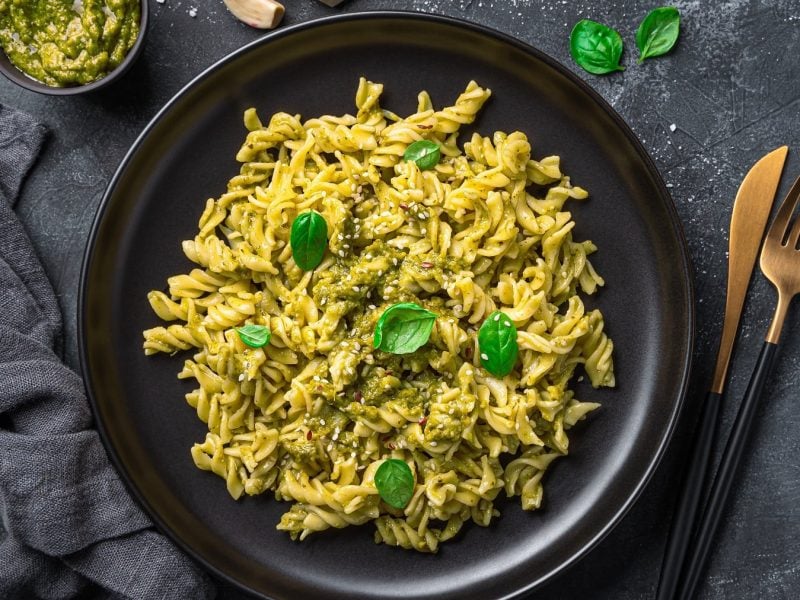 Ein schwarzer Teller mit dem Pasta-Gericht und schwarz-goldenem Besteck sowie grauem Tuch und Schale mit dem Pesto. Alles auf dunklem Untergrund von oben fotografiert.