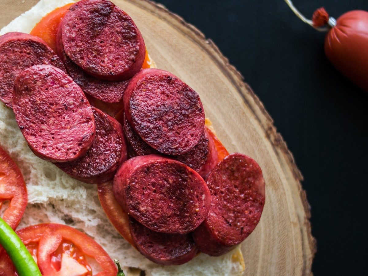 Draufsicht: eine aufgeklappte Sucuk-Stulle auf einem Holzbrett. Neben der Sucuk liegen auf dem Weißbrot eine Tomate und eine grüne Peperoni.