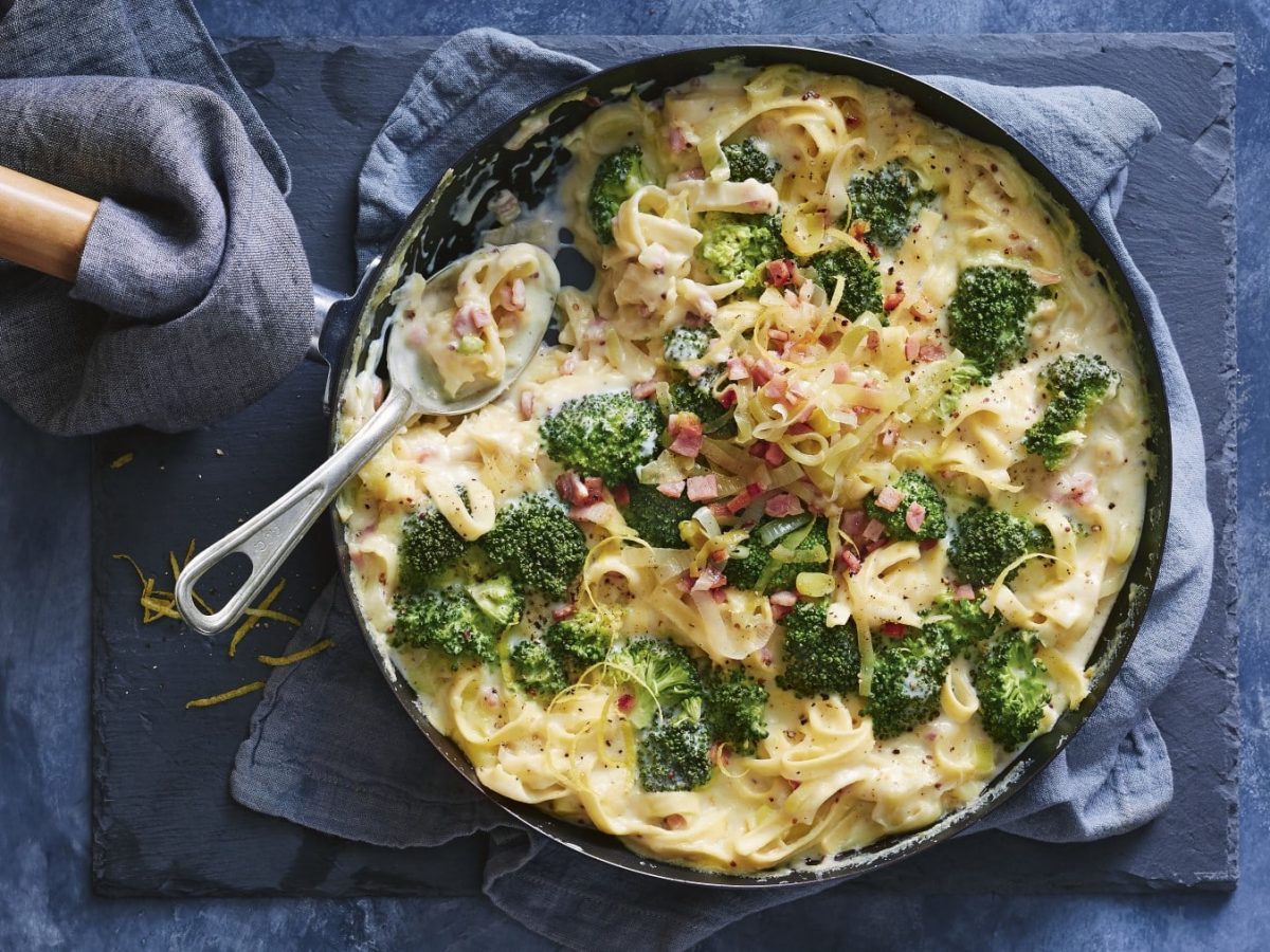 Tagaliatelle mit Käsesauce und Brokkoli in einer schwarzen Pfanne auf einem dunkelgrauen Geschirrtuch auf einer grauen Schieferplatte. Der Hintergrund ist ebenfalls dunkelgrau.