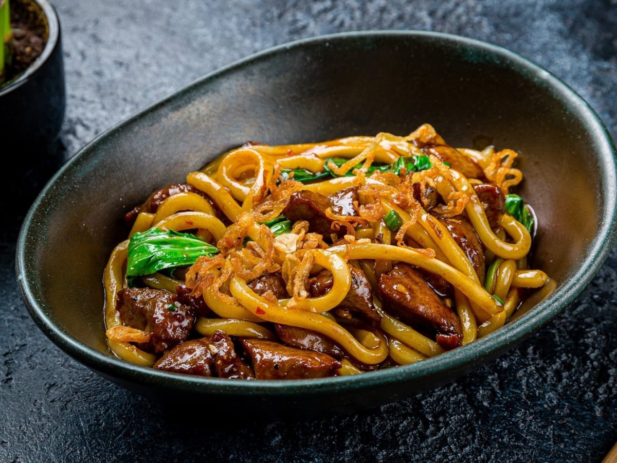 Eine schwarze Schale mit den Udon Nudeln auf einem dunklen Tisch. Daneben der untere Teil einer Bambus-Pflanze. Alles seitlich fotografiert.