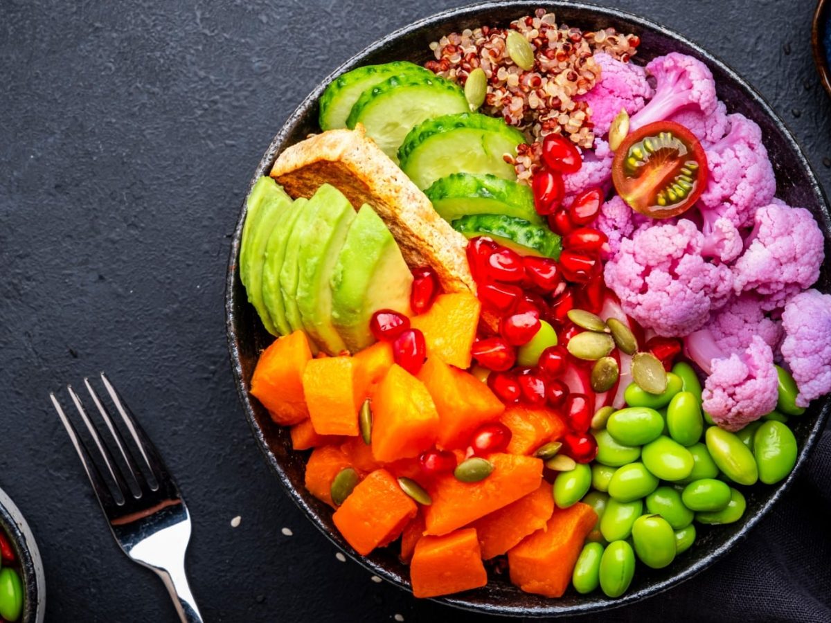 Vegane Regenbogen-Bowl auf schwarzem Untergrund, daneben Zutaten und eine Gabel.