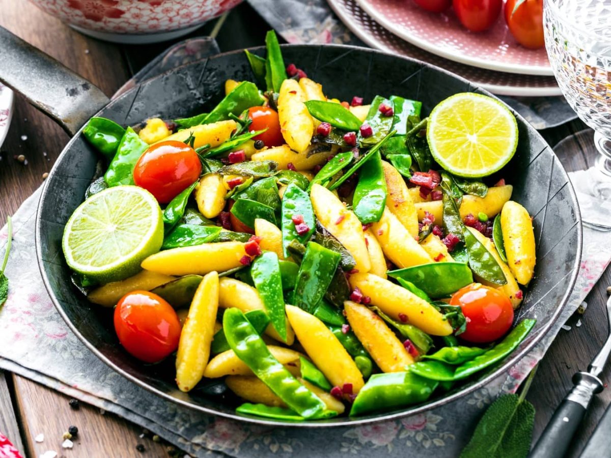 Vegane Schupfnudel-Pfanne mit Zuckerschoten, kleinen Tomaten, veganem Speck und Limette. Drum herum Zutaten und Geschirr.