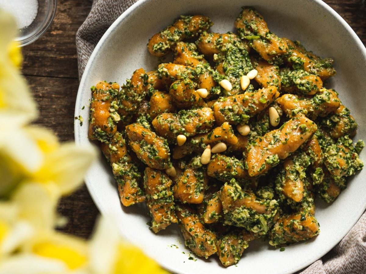 Draufsicht: Vegane Süßkartoffel-Gnocchi mit Spinat-Pesto und Pinienkernen auf weißem Teller mit Holzuntergrund und unscharfen gelben Pflanzen daneben.