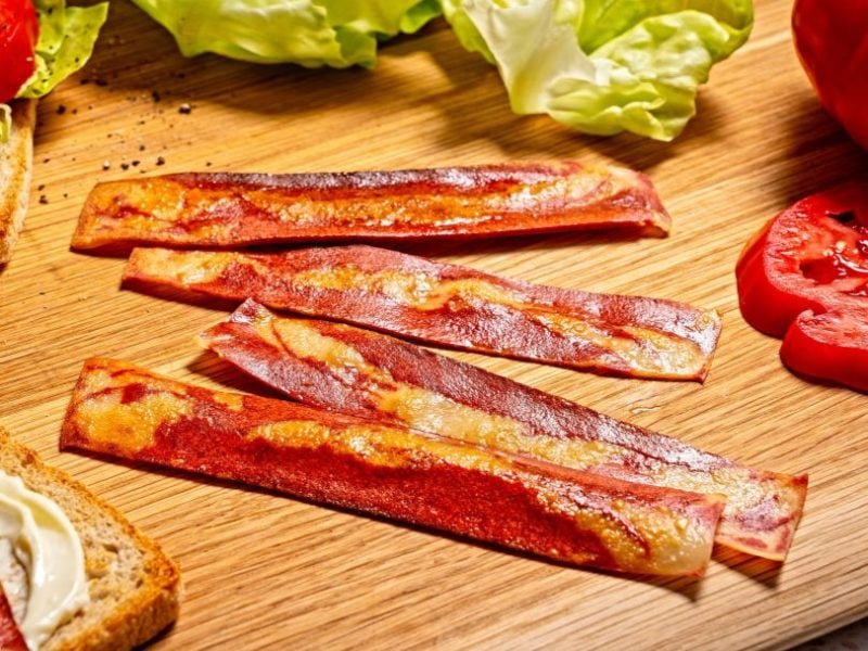 Die veganen Bacon-Streifen auf einem Holzschneidebrett mit Sandwichzutaten drumherum. Alles von schräg oben und recht nah fotografiert.