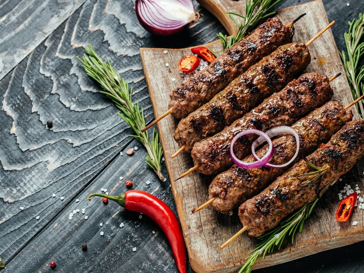 Mehrere Walnussköfte-Spieße auf einem Holzbrett von oben auf einem dunklen Tisch fotografiert. Daneben Rosmarinzweige, Chili und rote Zwiebel.