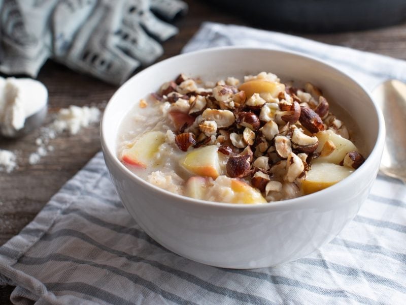 Draufsicht, schräg: Ein Apfel-Porridge mit gerösteten Nüssen. in einer Schüssel liegt auf einem Tuch, dass auf einem Holztisch liegt. Daneben sind ein Löffel, eine Hantel und ein Handschuh sowie ein weißes Pulver zu sehen.