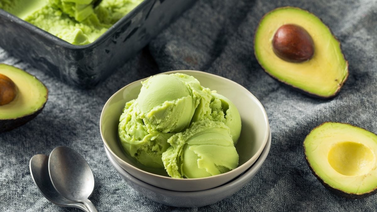 Drei Kugeln cremiges Avocado-Eis in einer grauen Schale, daneben frische Avocadohälften auf einer Stoffunterlage mit zwei Löffeln.