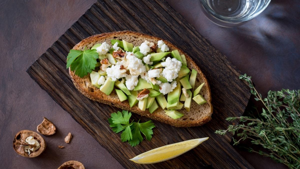 Draufsicht: Ein Avocado-Feta-Sandwich auf einem dunklem Brettchen aus Holz. Drumherum liegen weitere Lebensmittel.