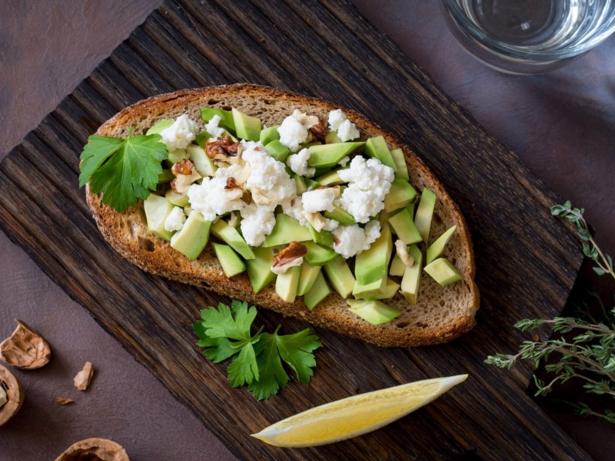 Draufsicht: Ein Avocado-Feta-Sandwich auf einem dunklem Brettchen aus Holz. Drumherum liegen weitere Lebensmittel.