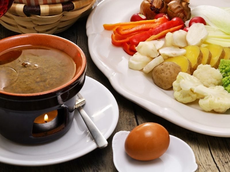 Bagna cauda in einem Stövchen. Daneben ein Ei und frisches Gemüse.