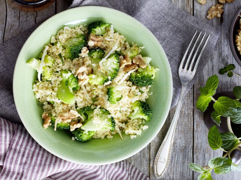 Bulgur mit Brokkoli in einer hellgrünen Schüssel auf einem grauen Geschirrtuch auf hölzernem Hintergrund. Rechts ein Schüsselchen mit Walnüssen und frische Minze.