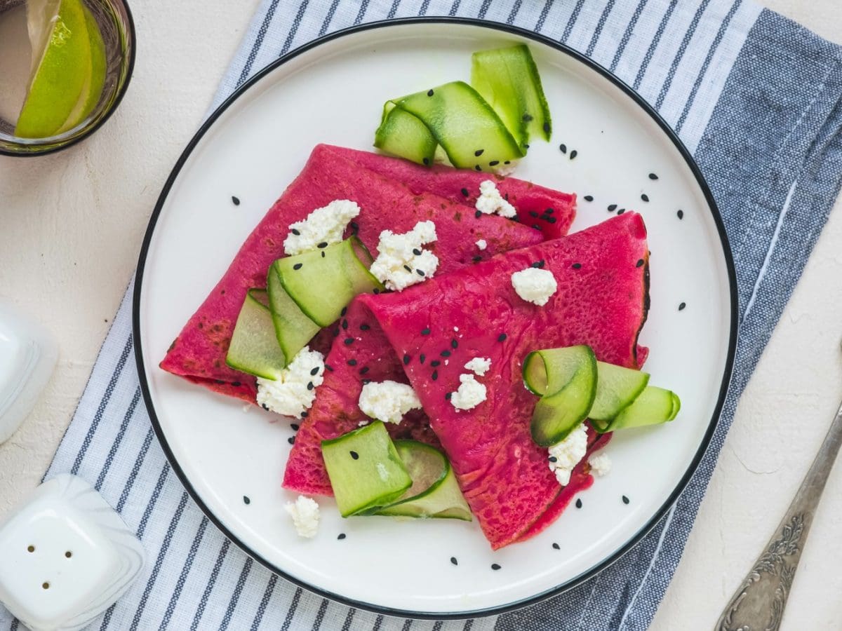 Rote Bete Crêpes mit Sesam, Hüttenkäse und Gurke auf einem weißen Teller auf einem blau-weiß-gestreiftem Geschirrtuch. Rechts davon eine Gabel, links auf dem Bild Salz- und Pfefferstreuer. Links oben ein Glas Wasser,