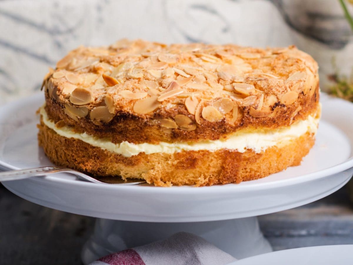 Veganer Bienenstich auf einem weißen Tortenreller. Der Hintergrund ist grau. Rechts auf dem Bild Blumen.