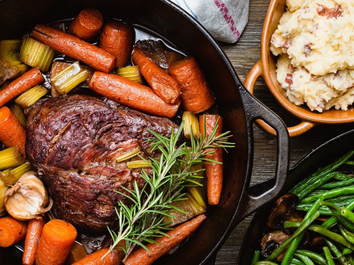 Draufsicht: Ein schwarzer gusseiserner Topf, in dem ein Braten und Schmorgemüse in einer Flüssigkeit ist, steht auf einem Untergrund aus Holz. Es handelt sich um einen italienischen Brasato al Barolo.