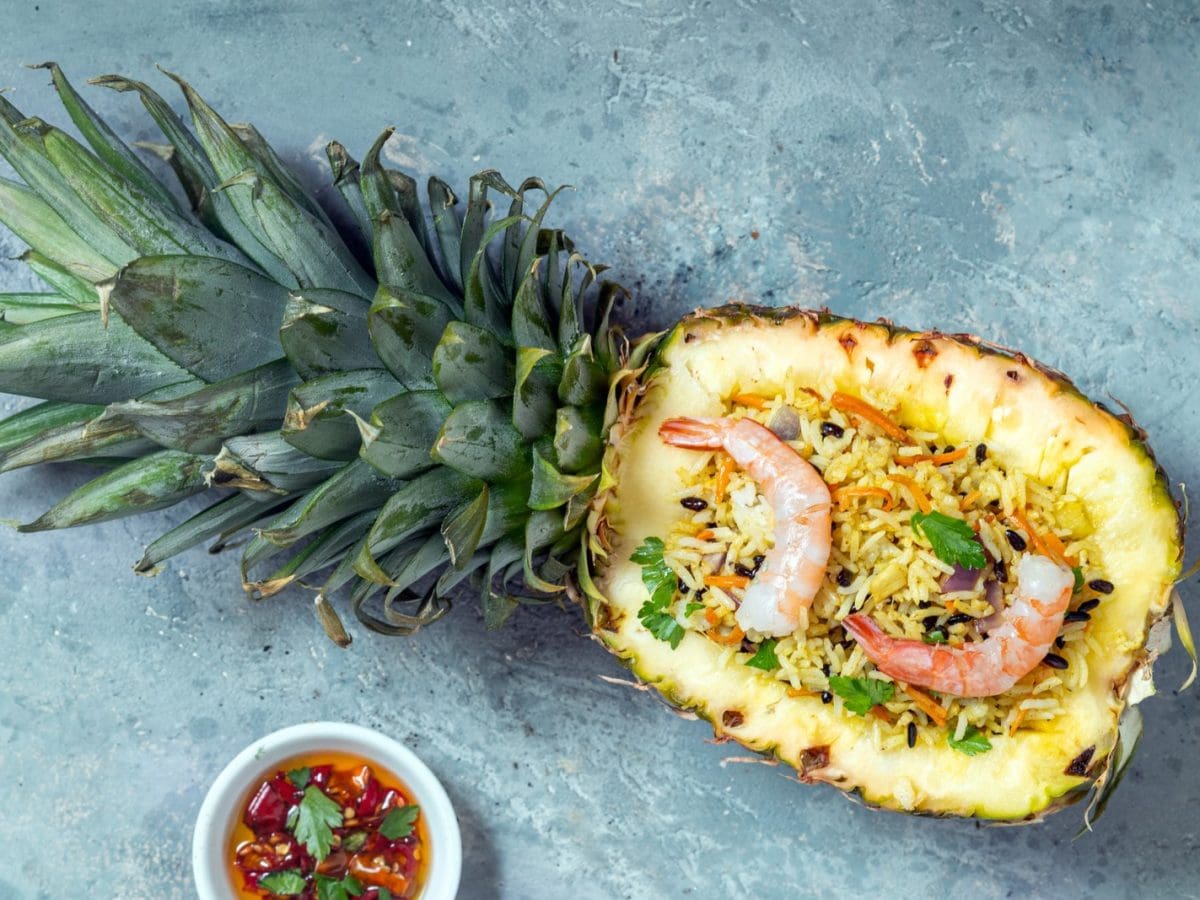 Eine ausgehöhlte Ananas mit dem Reisgericht auf einem hellblauen Tisch. Alles direkt von oben fotografiert.