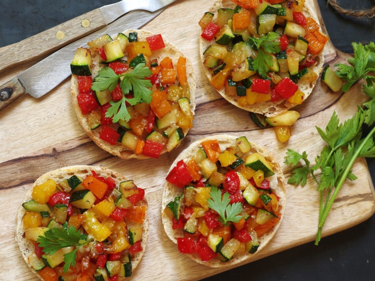 Ratatouille-Brötchen auf einem hölzernen Schneidebrett. Links Besteck. Der Hintergrund ist dunkelgrau.