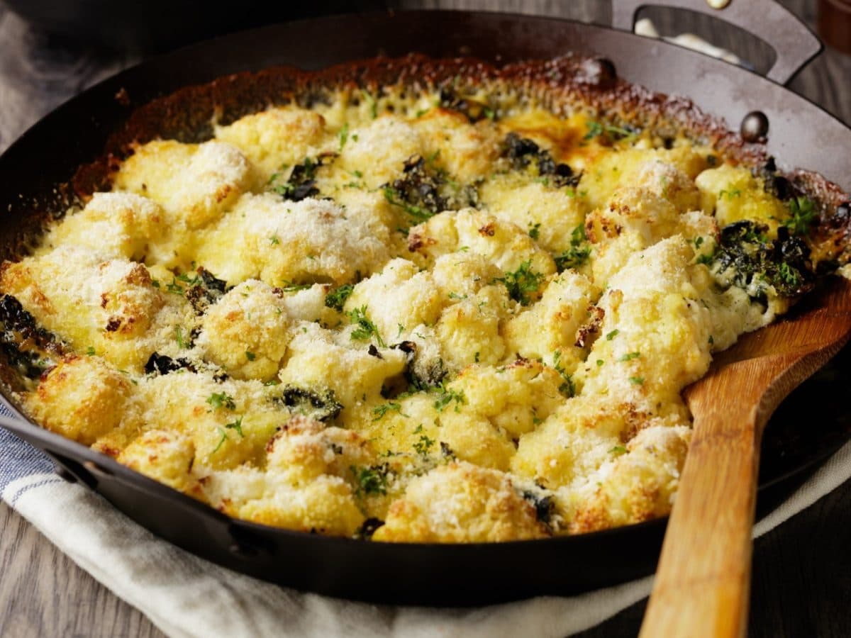 Draufsicht, schräg: Ein Blumenkohl-Grünkohl-Gratin in einer gusseisernen Pfanne. Die steht auf einem Tuch, dass auf einem Untergrund aus Holz ist. In der Pfanne ist ein großer Löffel aus Holz.