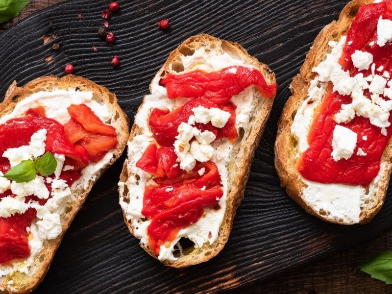 Draufsicht: drei Bruschetta mit gerösteter Paprika und Ricotta auf schwarzem Untergrund. Drum herum liegen frische Basilikumblätter.