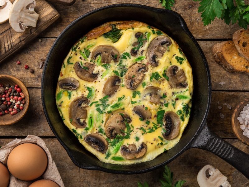 Draufsicht: Eine dunkle Pfanne in der eine Champignon-Frittata ist. Die steht auf einem rustikalem Tisch aus Holz. Drum herum liegen viele Zutaten für das Rezept.