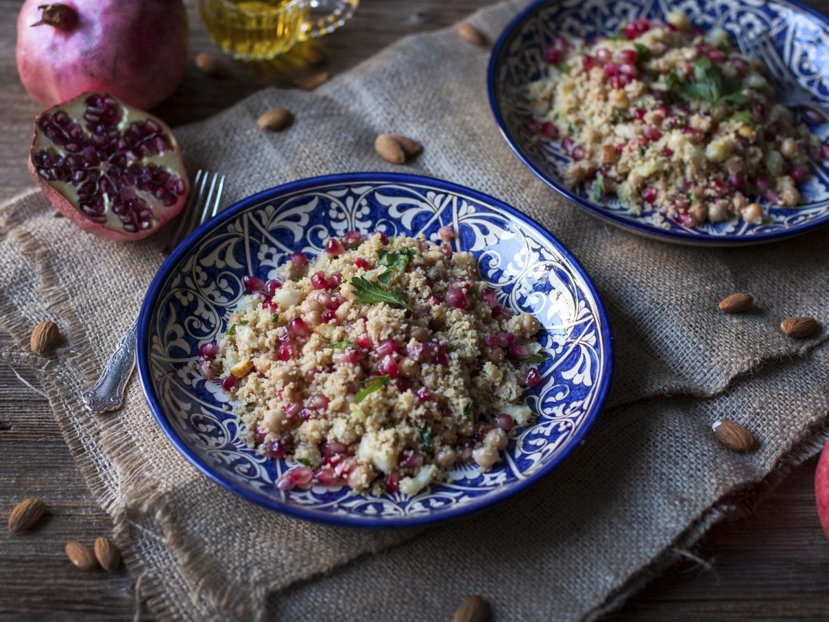 Draufsicht: Zwei Teller Couscous-Salat mit Granatapfel auf einem Untergrund aus Holz. Drum herum liegen Mandeln und ein aufgeschnittener Granatapfel.