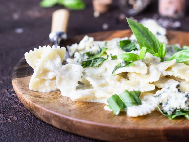 Frontal: Ein Holzteller mit Farfalle in Gorgonzola-Nuss-Sauce mit Basilikum. Im Hintergrund liegt frisches Basilikum.