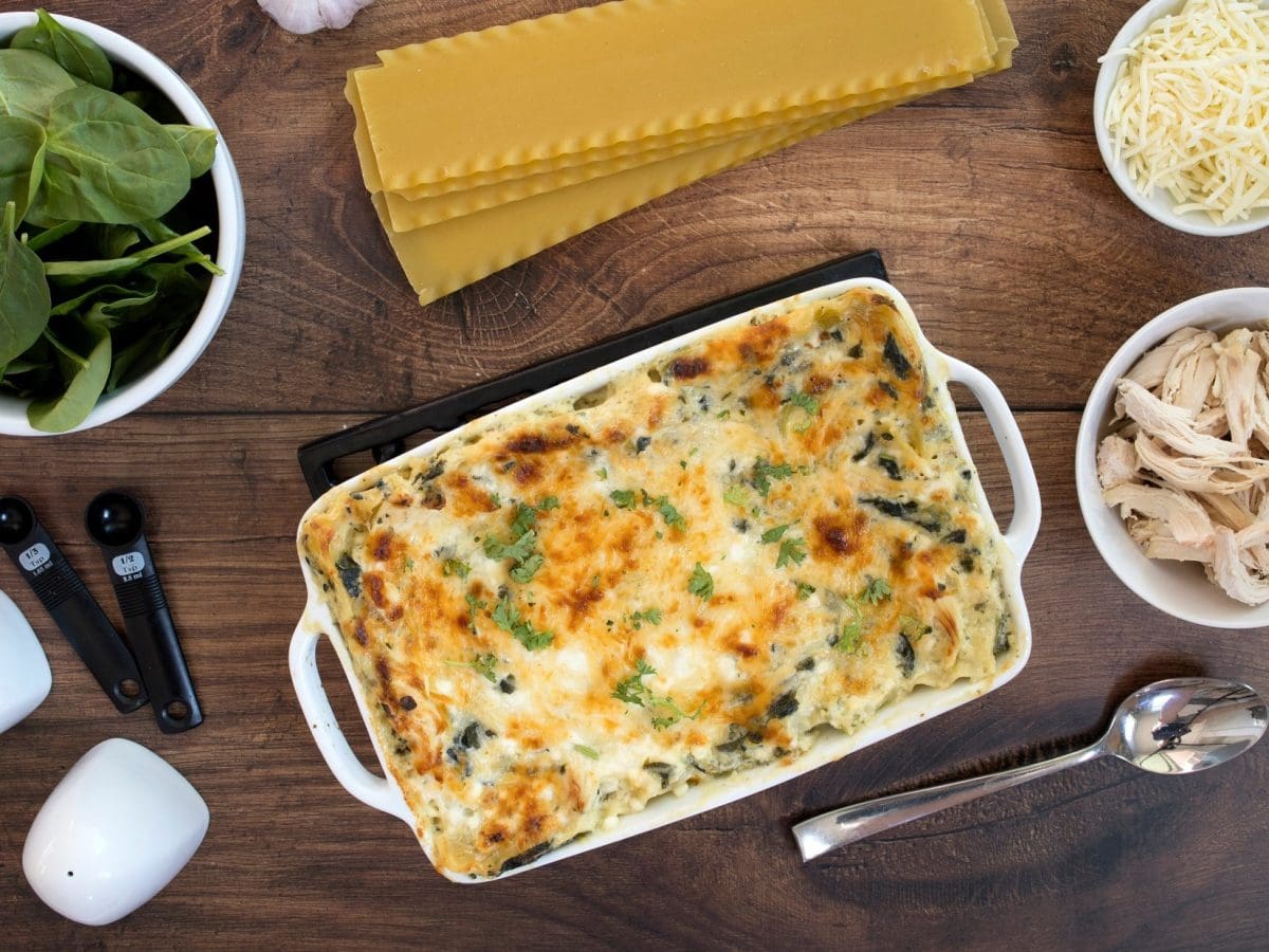 Auf einem dunklen Holztisch liegen die Zutaten sowie eine fertige Geflügel-Lasagne mit Spinat.