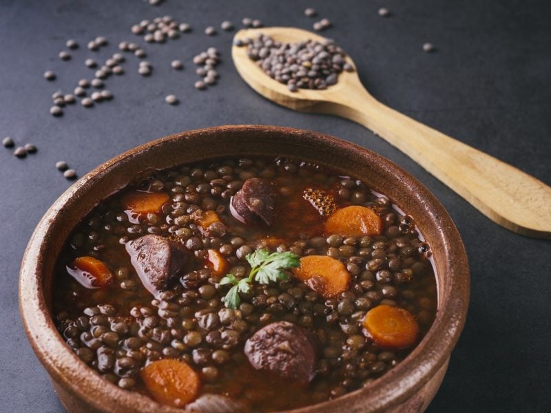 Draufsicht schräg: ein Linseneintopf mit Chorizo in einer Tonschale. Daneben liegen ein Holzlöffel und getrocknete braune Linsen.