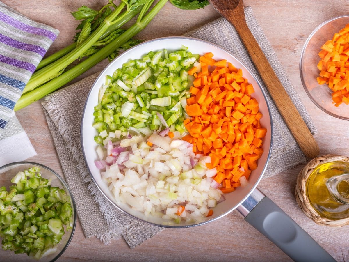 Draufsicht: Auf einer Arbeitsplatte in einer Küche liegt eine Pfanne mit gleichmäßig geschnittenem Gemüse. Es handelt sich um ein Mirepoix. Drum herum liegen Küchenhandtücher und klein geschnittenes Gemüse.