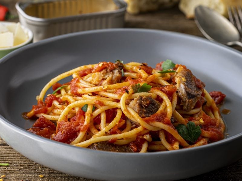 Frontal: Ein Teller Pasta mit Sardinien. auf einem Holztisch. Im Hintergrund sind Butter, Weißbrot und einen Dose, in der die Sardinen waren.