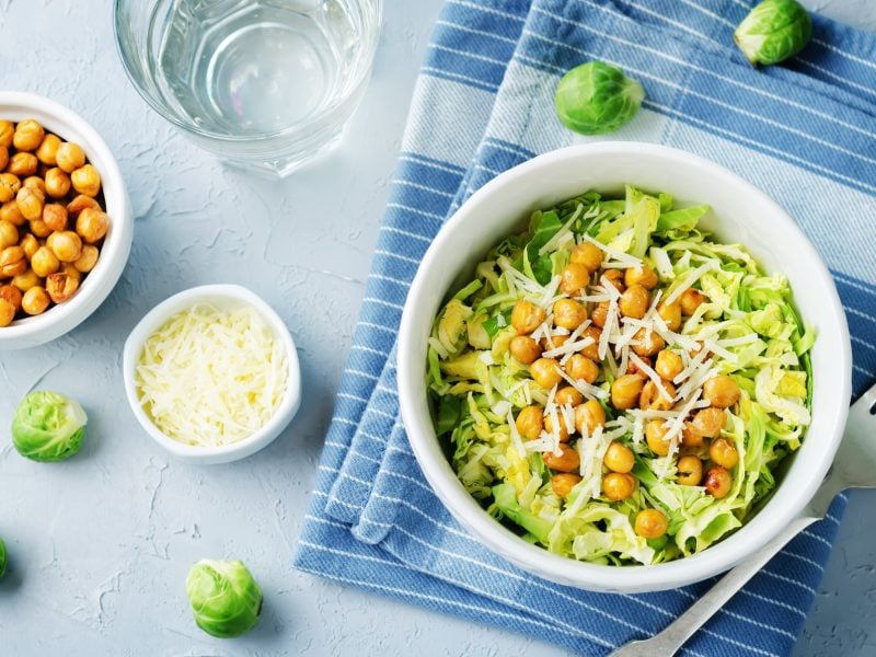 Draufsicht: auf hellblauem Untergrund ligt ein hellblaues Tuche auf dem eine Schale mit Rosenkohl-Kichererbsen-Salat steht. Drum herum sind eine Schale mit Kichererbsen, eine Schale mit geriebenem Käse sowie roher Rosenkohl.