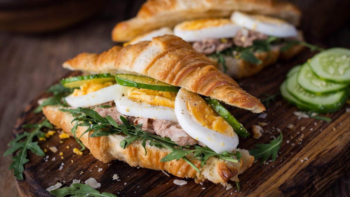 Draufsicht, schräg: Auf einem Holzbrett liegen zwei Thunfisch-Croissant mit Ei. Daneben liegen einige Scheiben Salatgurke.