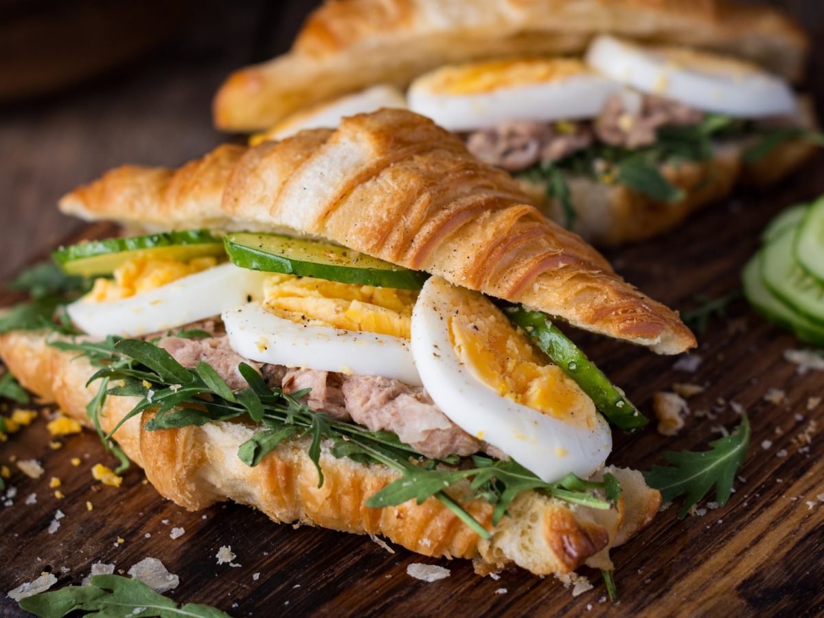 Draufsicht, schräg: Auf einem Holzbrett liegen zwei Thunfisch-Croissant mit Ei. Daneben liegen einige Scheiben Salatgurke.