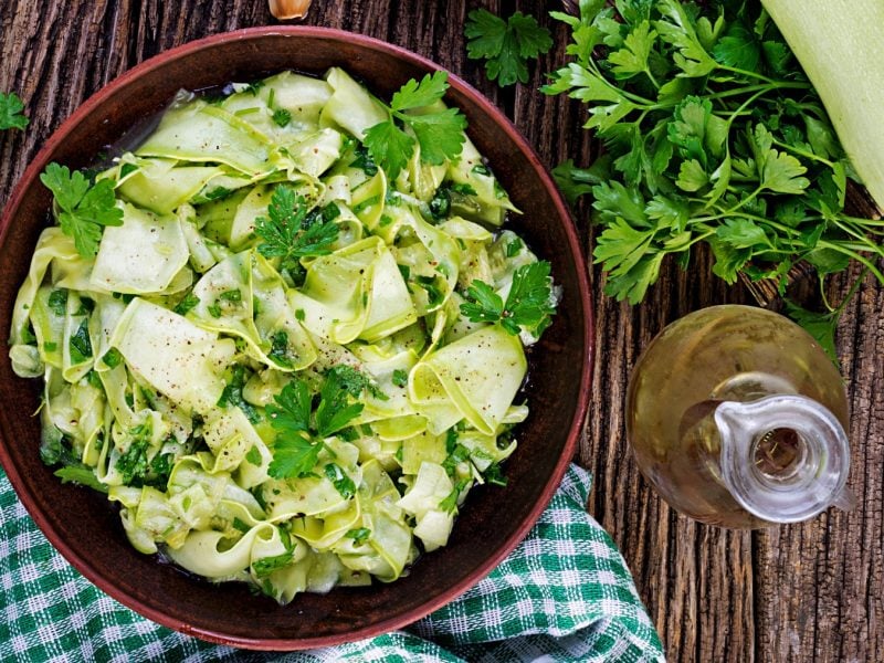 Eingelegter Zucchini-Salat in einer Schüssel mit Petersilie darauf und daneben auf Holzuntergrund. Daneben Ölflasche, Küchentuch, Petersilie und eine helle Zucchini.