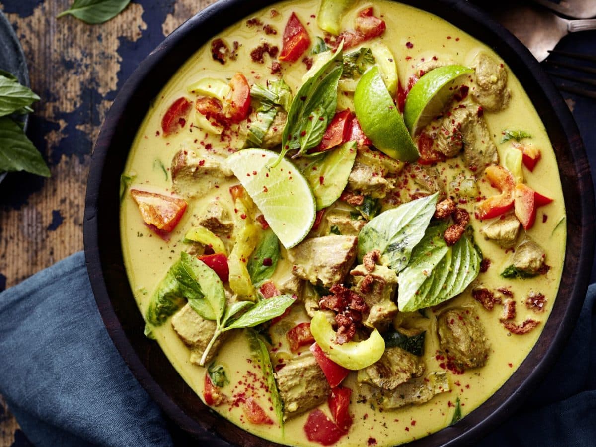 Entencurry in einer dunkelbraunen Schüssel, links davon Basilikum, rechts auf dem Bild Besteck. Der Hintergrund ist hölzern. Unten auf dem Bild ein dunkelgraues Geschirrtuch.