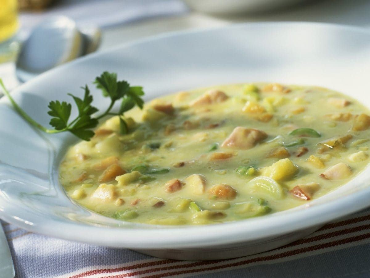 Erbsensuppe mit Würstchen in einem weißen Suppenteller auf einem Tisch. Links davon ein Löffel.