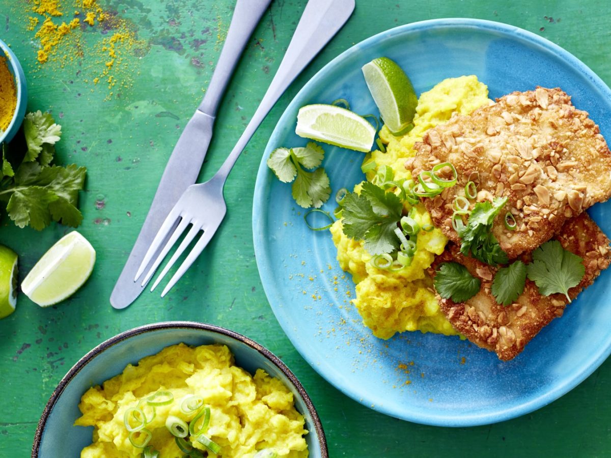 Erdnuss-Schnitzel mit Curry-Stampf auf einem blauen Teller auf grünem Hintergrund. Links davon Messer, Gabel, Curry und Limette. Unten auf dem Bild eine Schüssel mit Kartoffelstampf.