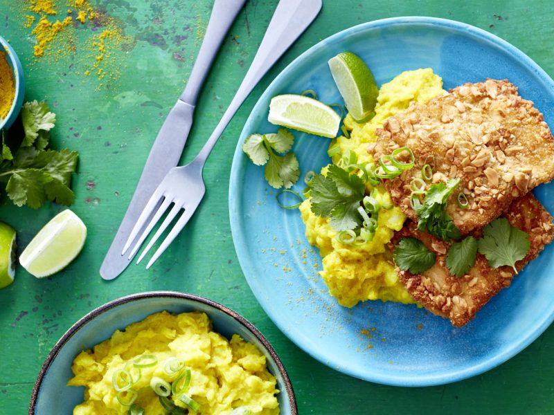 Erdnuss-Schnitzel mit Curry-Stampf auf einem blauen Teller auf grünem Hintergrund. Links davon Messer, Gabel, Curry und Limette. Unten auf dem Bild eine Schüssel mit Kartoffelstampf.