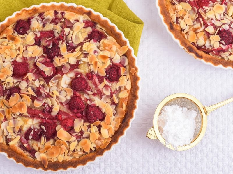 Frangipane-Tarte mit Rhabarber und Himbeeren. Daneben in goldenes Sieb mit Puderzucker.