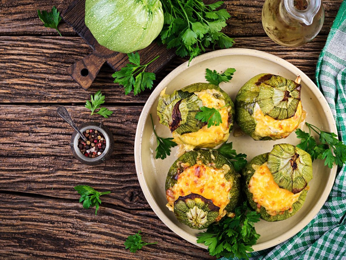 Vier gefüllte Zucchini mit Soja-Hack und Käse und Petersilie auf einem Teller, Holzuntergrund und Zutaten drum herum.