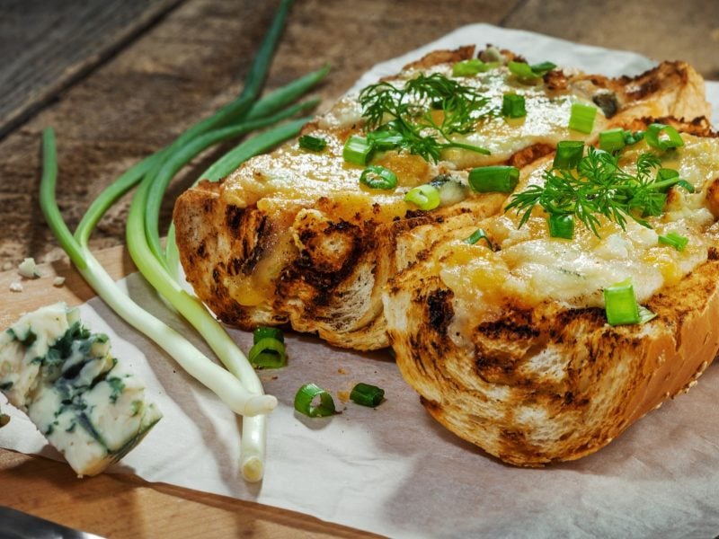 Draufsicht, schräg: Zwei halbierte Gorgonzola-Mozzarella-Baguette auf hellem Untergrund, der auf Holz liegt. Daneben liegen Blauschimmelkäsestücke sowie Frühlingszwiebeln und Kräuter.