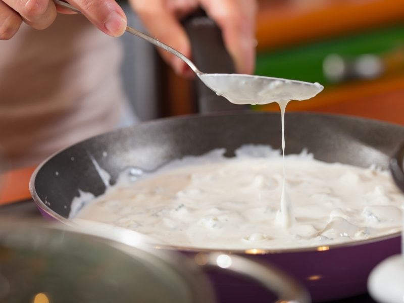 Frontalansicht: Eine Hand bereitet in einer Pfanne eine Gorgonzola-Sauce zu.