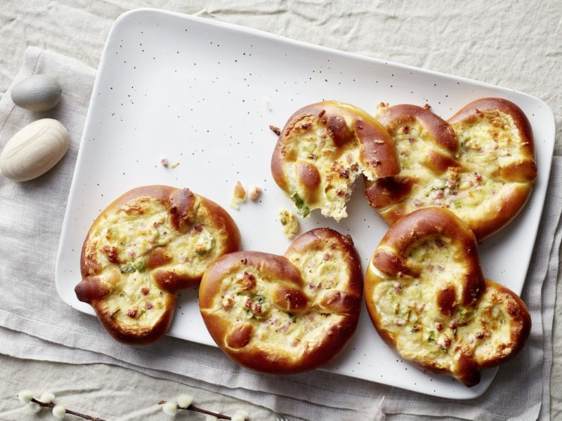 Überbackene Käsebrezeln Flammkuchenstyle auf einem weißen Teller mit hellem Stoffuntergrund.