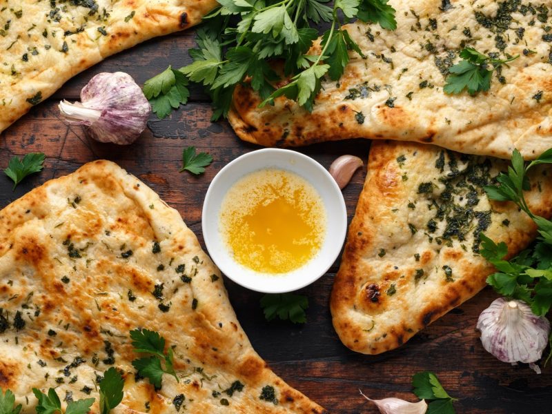 Knoblauch-Naan auf braunem Hintergrund verteilt. In der Mitte eine kleine Schüssel mit Knoblauchbutter. Knoblauch auf dem Bild verteilt.