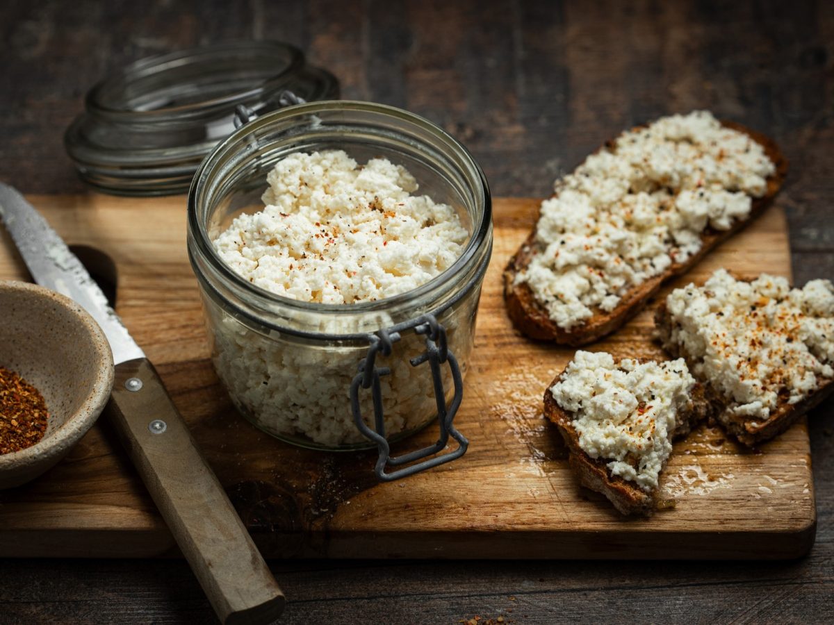 Ein dunkles Holzbrett auf dunklem Tisch mit einem Weckglas voll mit körnigem Frischkäse und beschmierten Broten. Daneben eine Schale rotes Gewürz und ein Messer. Alles von schräg oben fotografiert.