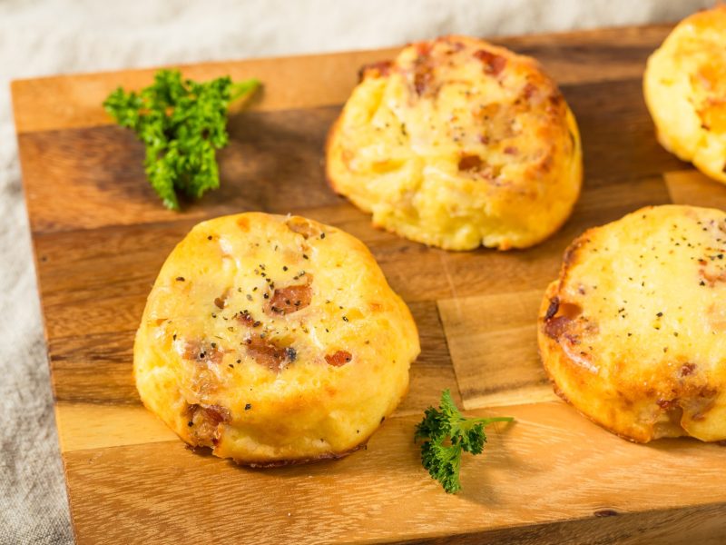 Ein Holzbrett mit vier kleinen Kartoffel-Soufflés auf hellem Untergrund. Alles von schräg oben fotografiert.