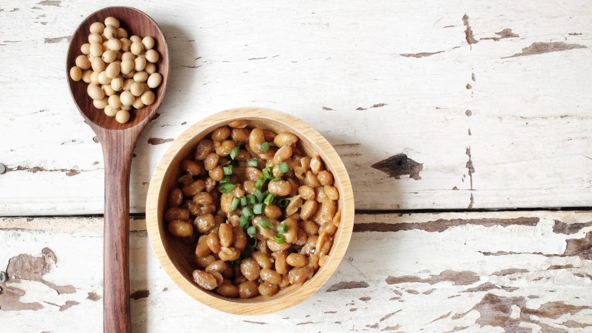 Natto in einer weißen Schüssel auf weißem Hintergrund. Links auf dem Foto ein Löffel mit Sojabohnen.